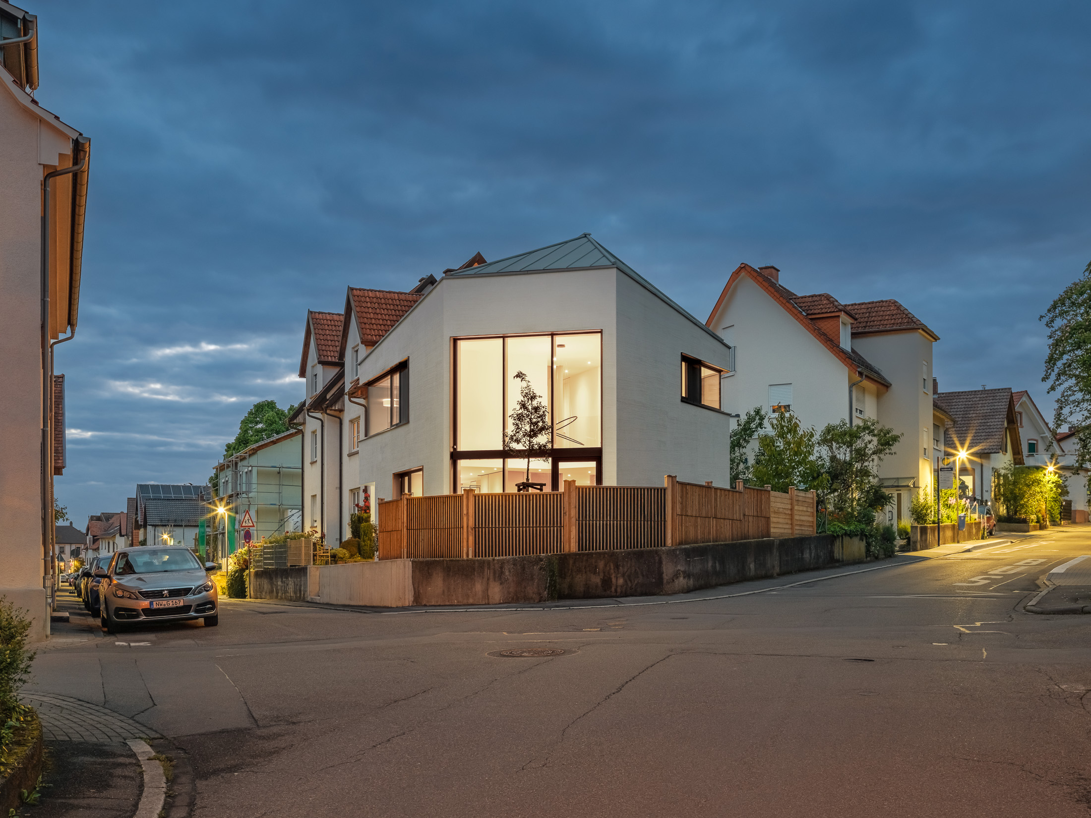 Monolithisches, kompaktes Wohnhaus mit polygonalem Grundriss und asymmetrischem Walmdach aus anthrazitfarbenem Stehfalzblech an einer markanten Verkehrskreuzung.