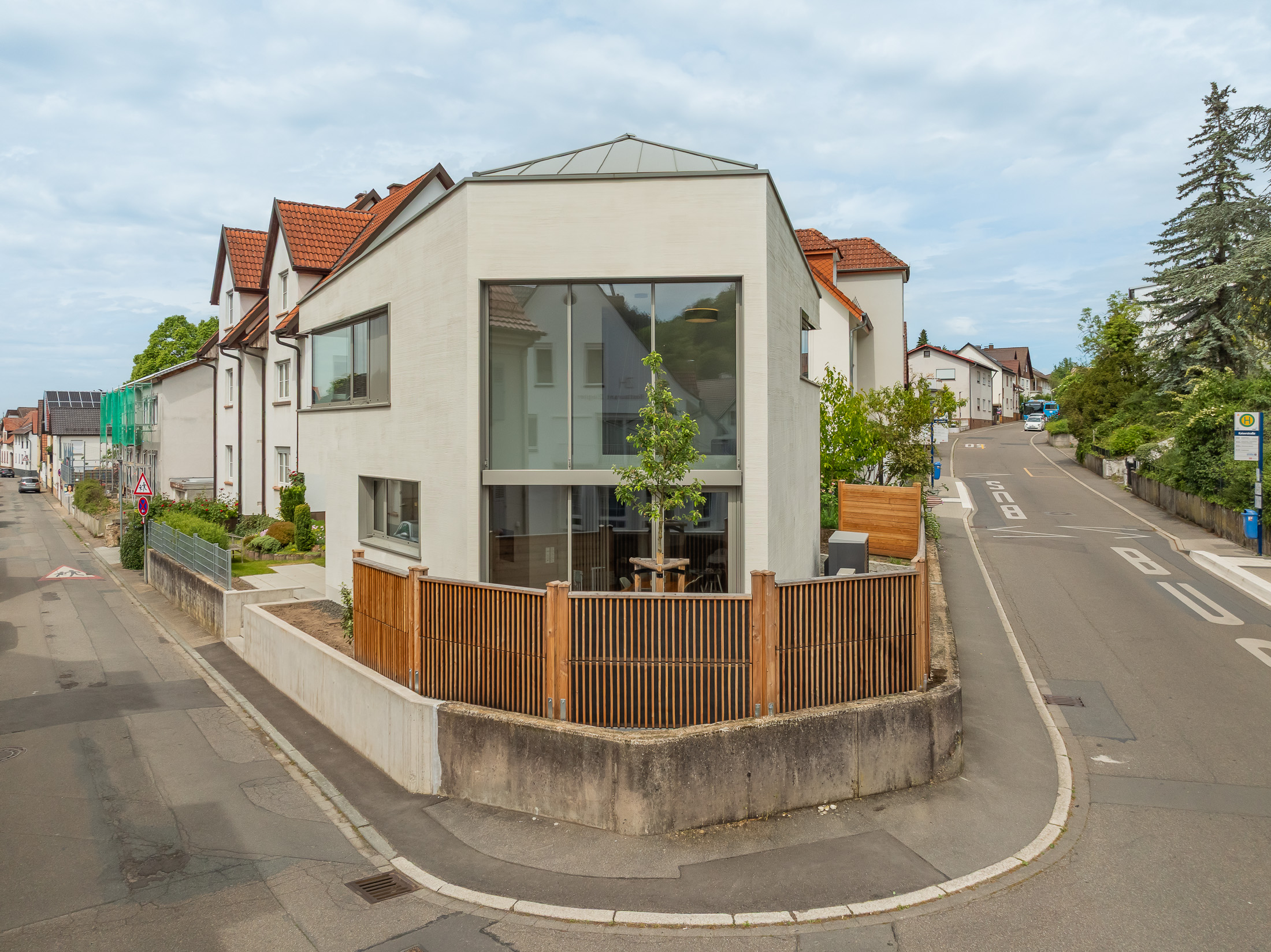 Monolithisches, kompaktes Wohnhaus mit polygonalem Grundriss und asymmetrischem Walmdach aus anthrazitfarbenem Stehfalzblech an einer markanten Verkehrskreuzung.