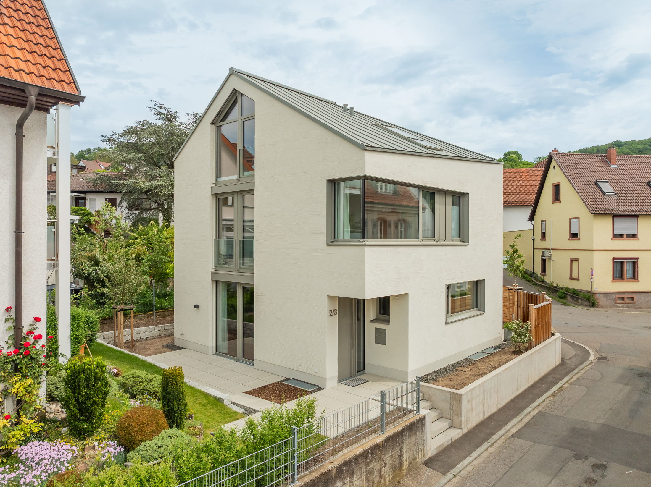 Monolithisches, kompaktes Wohnhaus mit polygonalem Grundriss und asymmetrischem Walmdach aus anthrazitfarbenem Stehfalzblech an einer markanten Verkehrskreuzung.