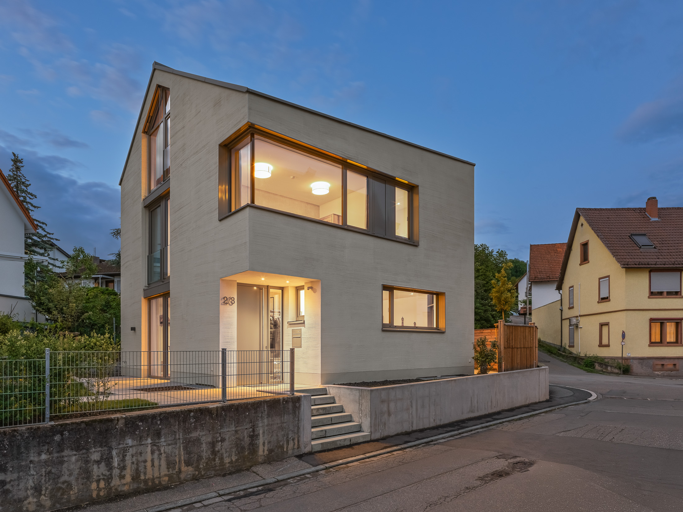 Monolithisches, kompaktes Wohnhaus mit polygonalem Grundriss und asymmetrischem Walmdach aus anthrazitfarbenem Stehfalzblech an einer markanten Verkehrskreuzung.