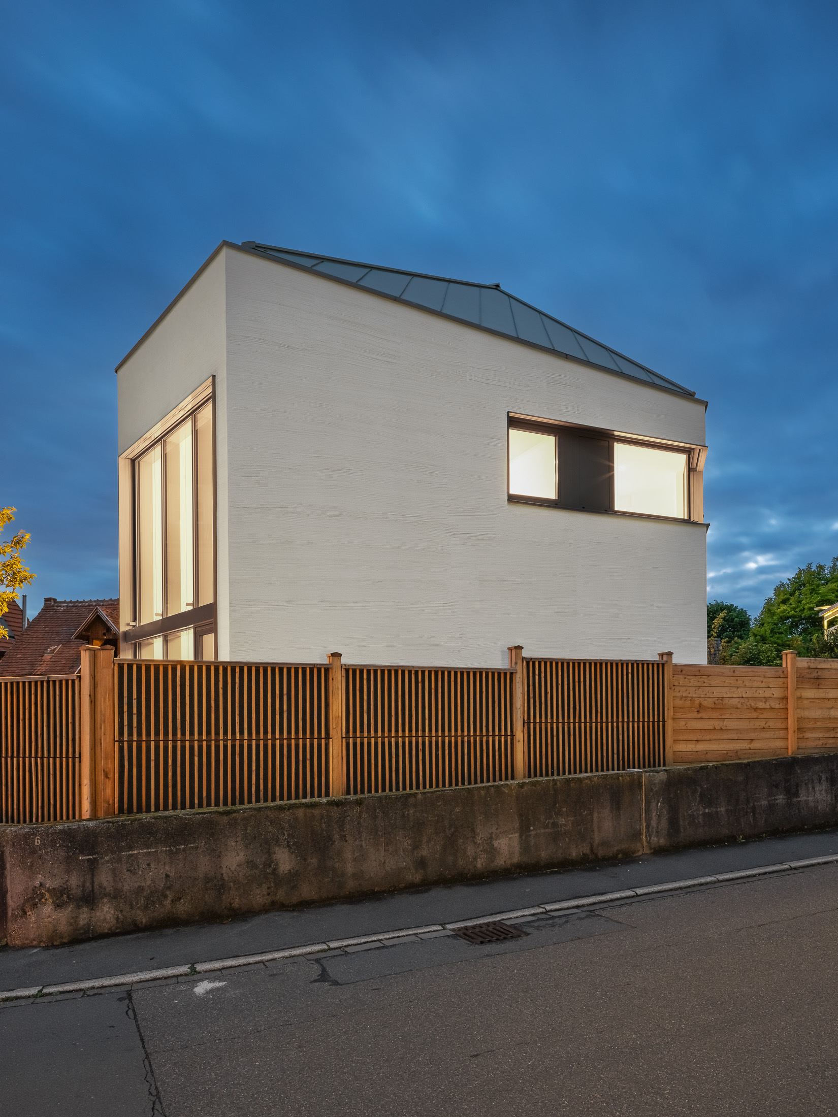 Monolithisches, kompaktes Wohnhaus mit polygonalem Grundriss und asymmetrischem Walmdach aus anthrazitfarbenem Stehfalzblech an einer markanten Verkehrskreuzung.
