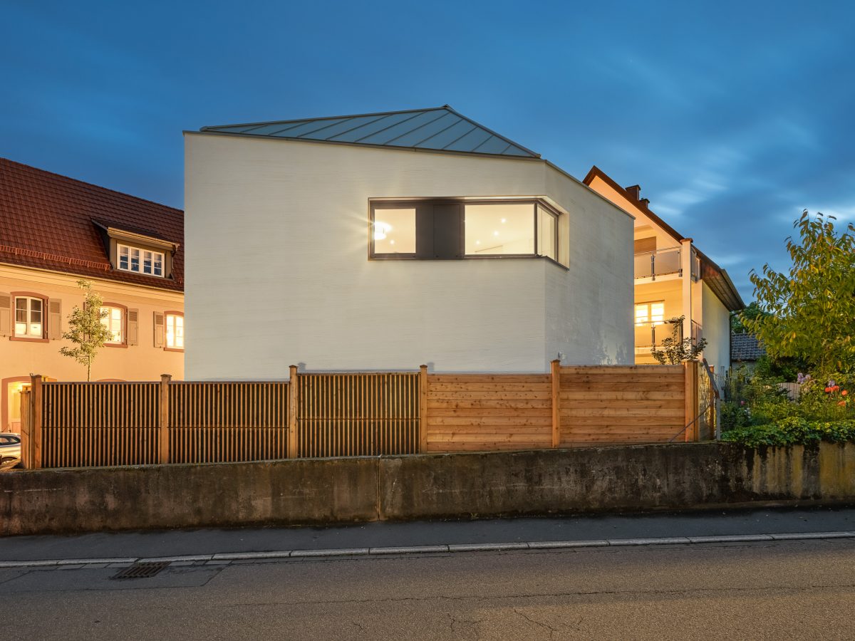 Monolithisches, kompaktes Wohnhaus mit polygonalem Grundriss und asymmetrischem Walmdach aus anthrazitfarbenem Stehfalzblech an einer markanten Verkehrskreuzung.