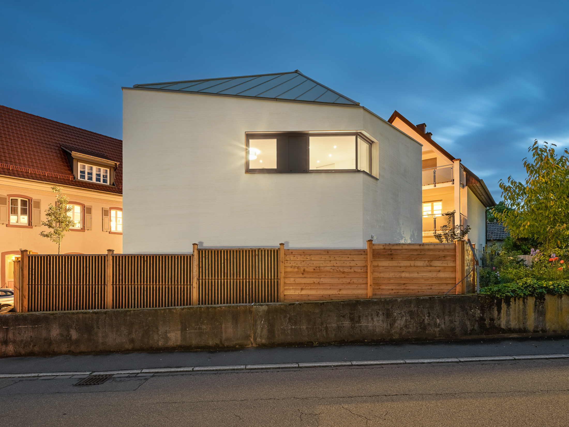 Monolithisches, kompaktes Wohnhaus mit polygonalem Grundriss und asymmetrischem Walmdach aus anthrazitfarbenem Stehfalzblech an einer markanten Verkehrskreuzung.
