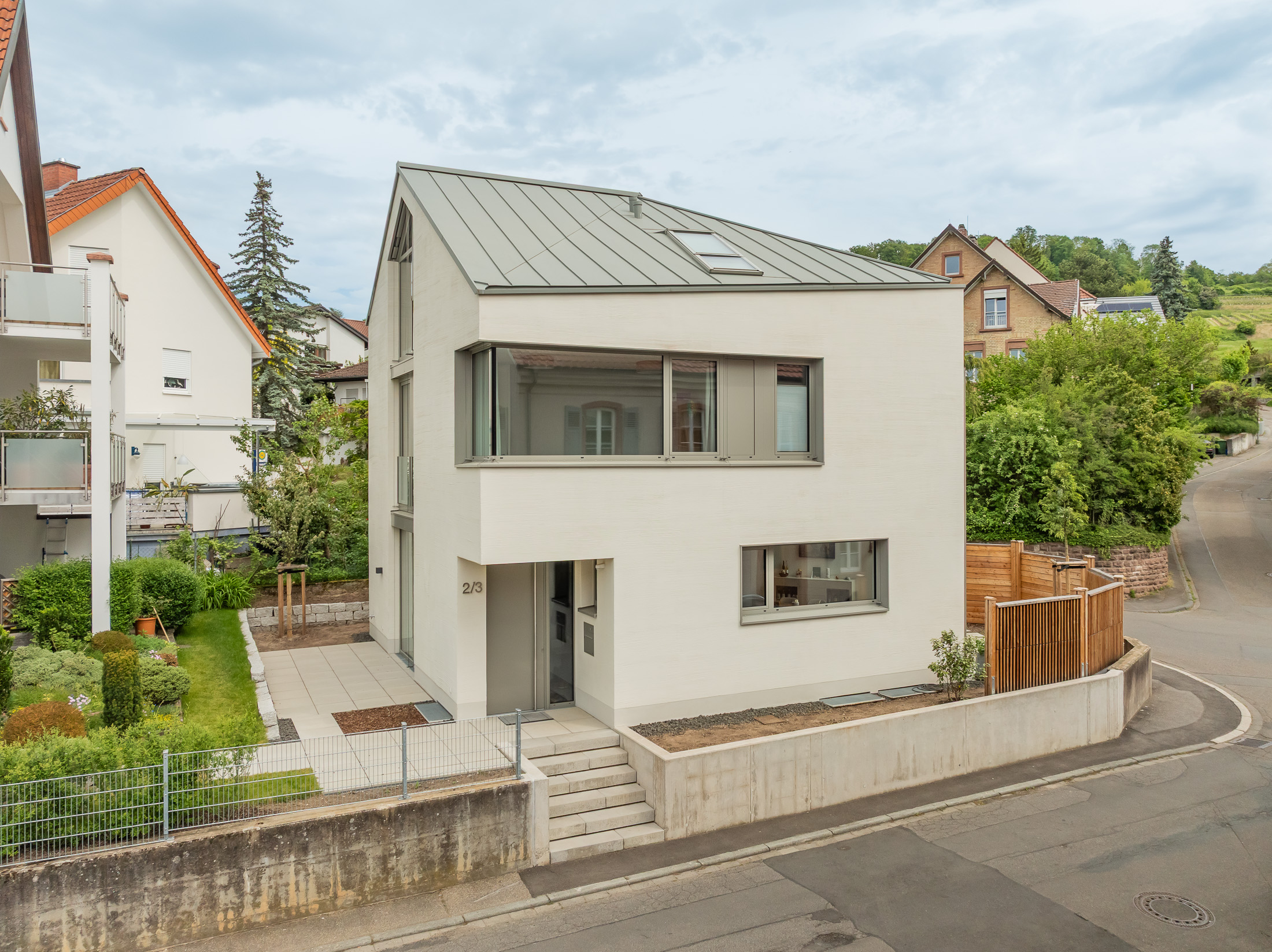 Monolithisches, kompaktes Wohnhaus mit polygonalem Grundriss und asymmetrischem Walmdach aus anthrazitfarbenem Stehfalzblech an einer markanten Verkehrskreuzung.