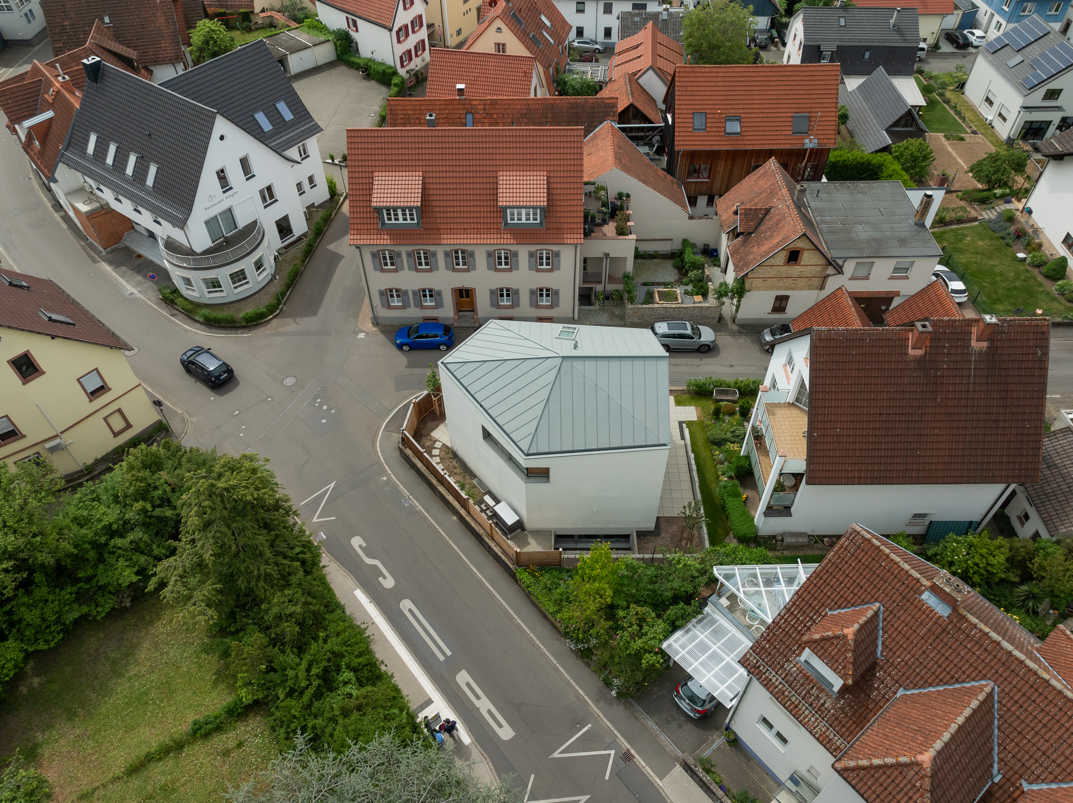 Monolithisches, kompaktes Wohnhaus mit polygonalem Grundriss und asymmetrischem Walmdach aus anthrazitfarbenem Stehfalzblech an einer markanten Verkehrskreuzung.