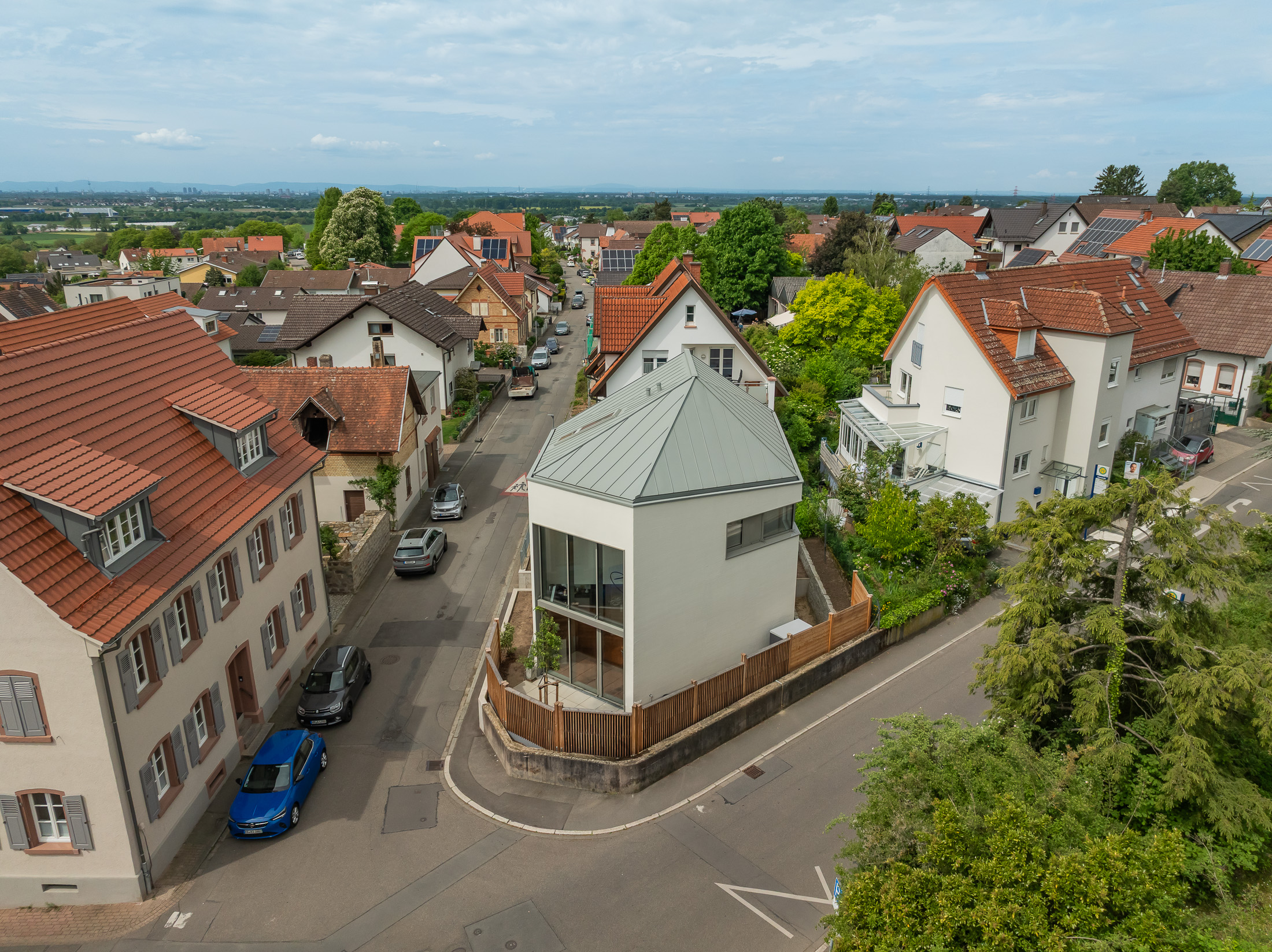 Monolithisches, kompaktes Wohnhaus mit polygonalem Grundriss und asymmetrischem Walmdach aus anthrazitfarbenem Stehfalzblech an einer markanten Verkehrskreuzung.