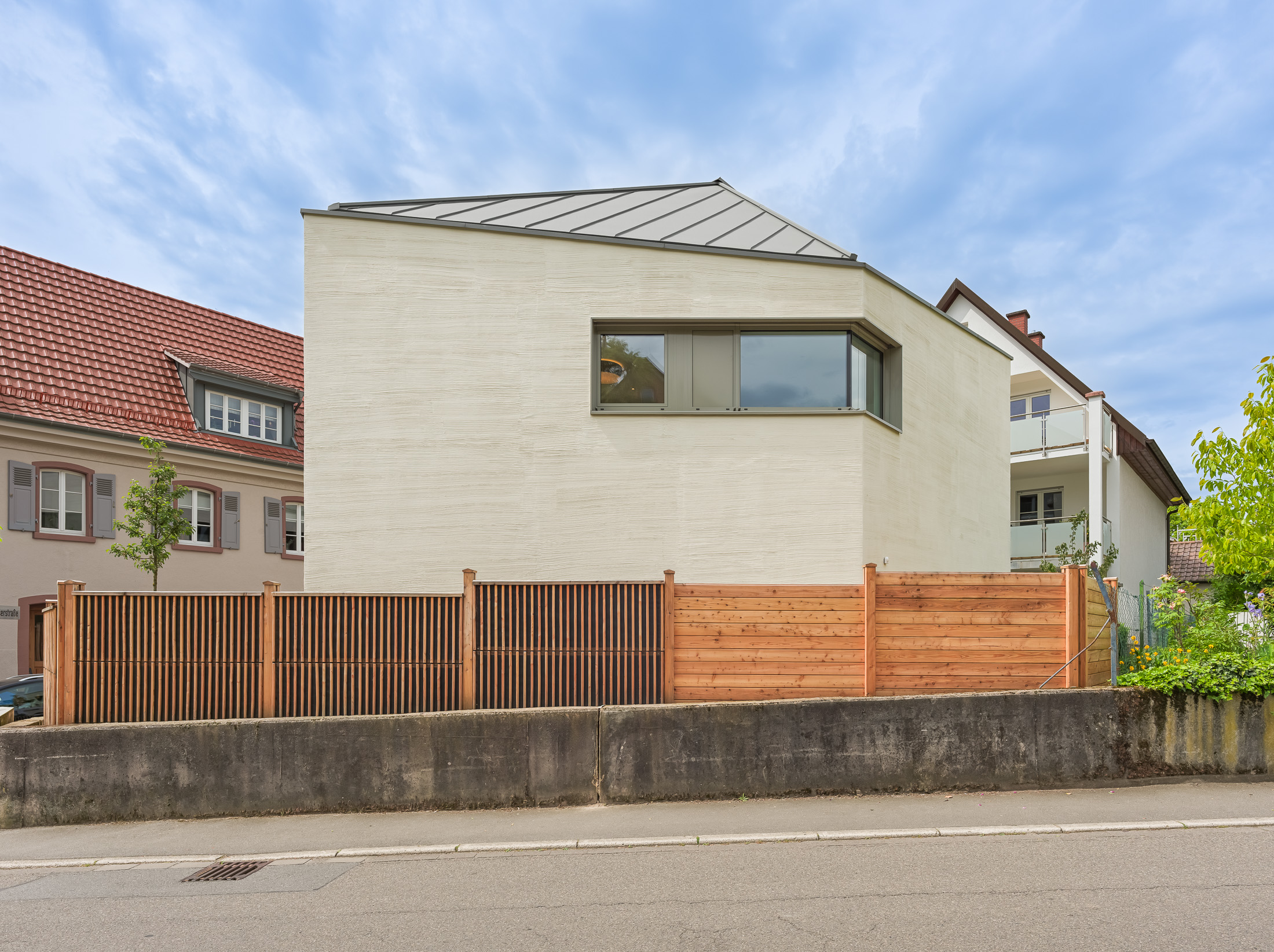 Monolithisches, kompaktes Wohnhaus mit polygonalem Grundriss und asymmetrischem Walmdach aus anthrazitfarbenem Stehfalzblech an einer markanten Verkehrskreuzung.