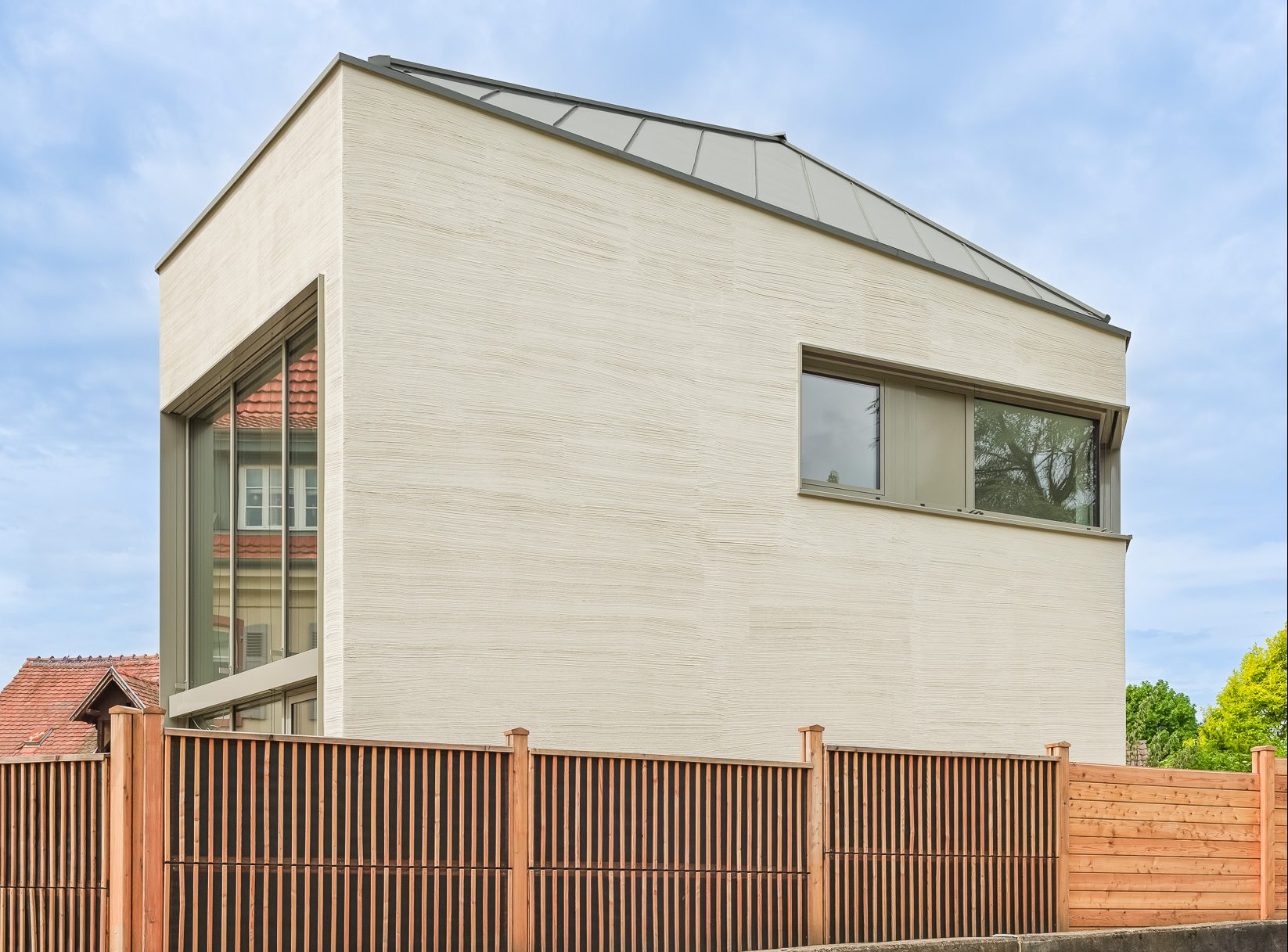Monolithisches, kompaktes Wohnhaus mit polygonalem Grundriss und asymmetrischem Walmdach aus anthrazitfarbenem Stehfalzblech an einer markanten Verkehrskreuzung.