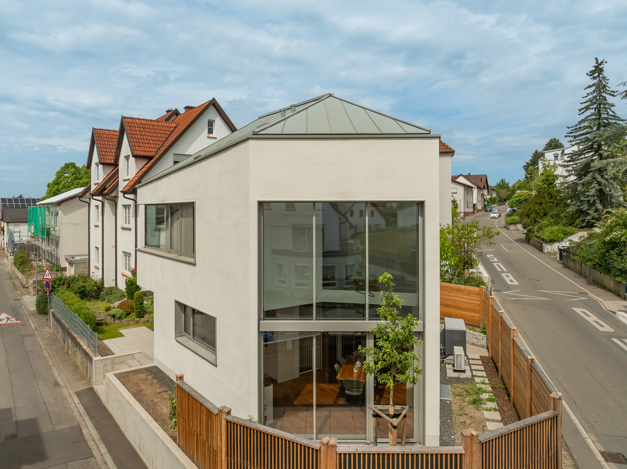 Monolithisches, kompaktes Wohnhaus mit polygonalem Grundriss und asymmetrischem Walmdach aus anthrazitfarbenem Stehfalzblech an einer markanten Verkehrskreuzung.