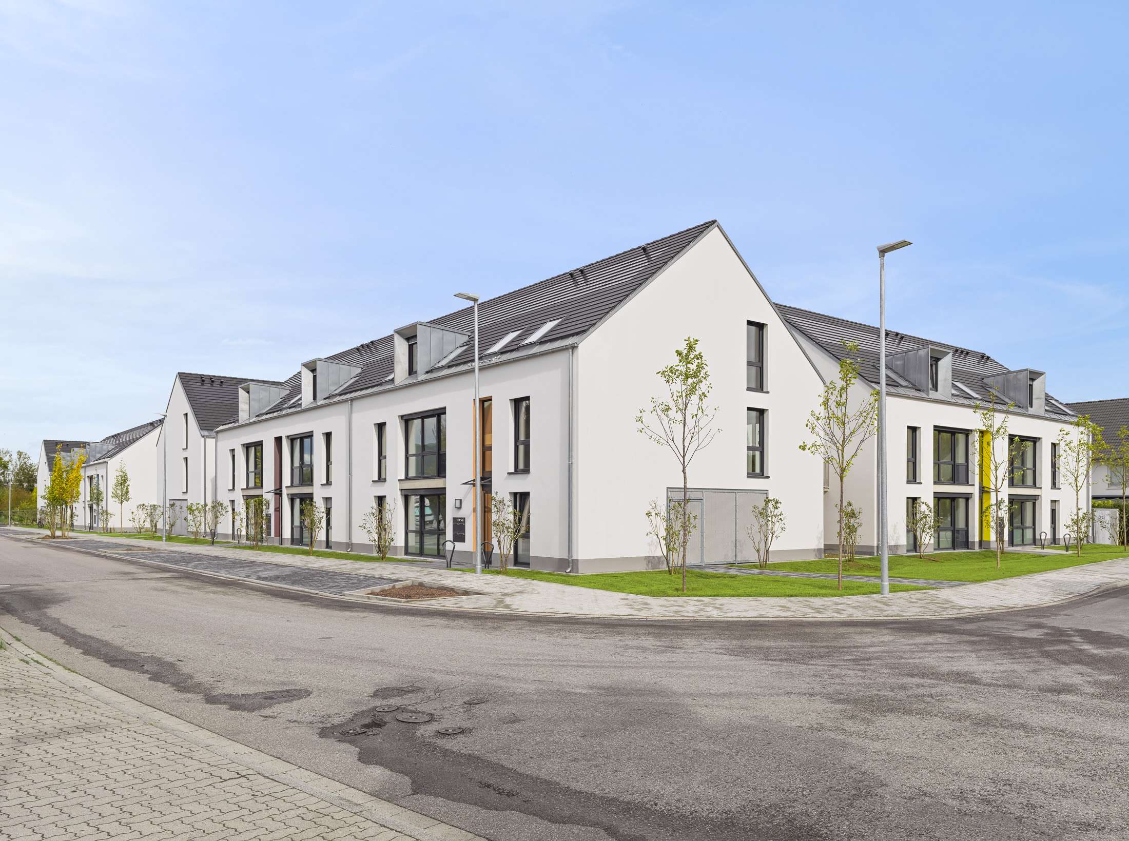 Mehrfamilienhausanlage für die VOLKSWOHNUNG GmbH in Karlsruhe-Durlach mit zweigeschossigen Hausgruppen und prägnanten Satteldächern, die sich um zwei begrünte, geschützte Innenhöfe organisieren; großzügige Verglasungen, Loggien und Dachgauben sowie eine flächenbündig integrierte Indach-Photovoltaikanlage prägen das architektonische Erscheinungsbild.