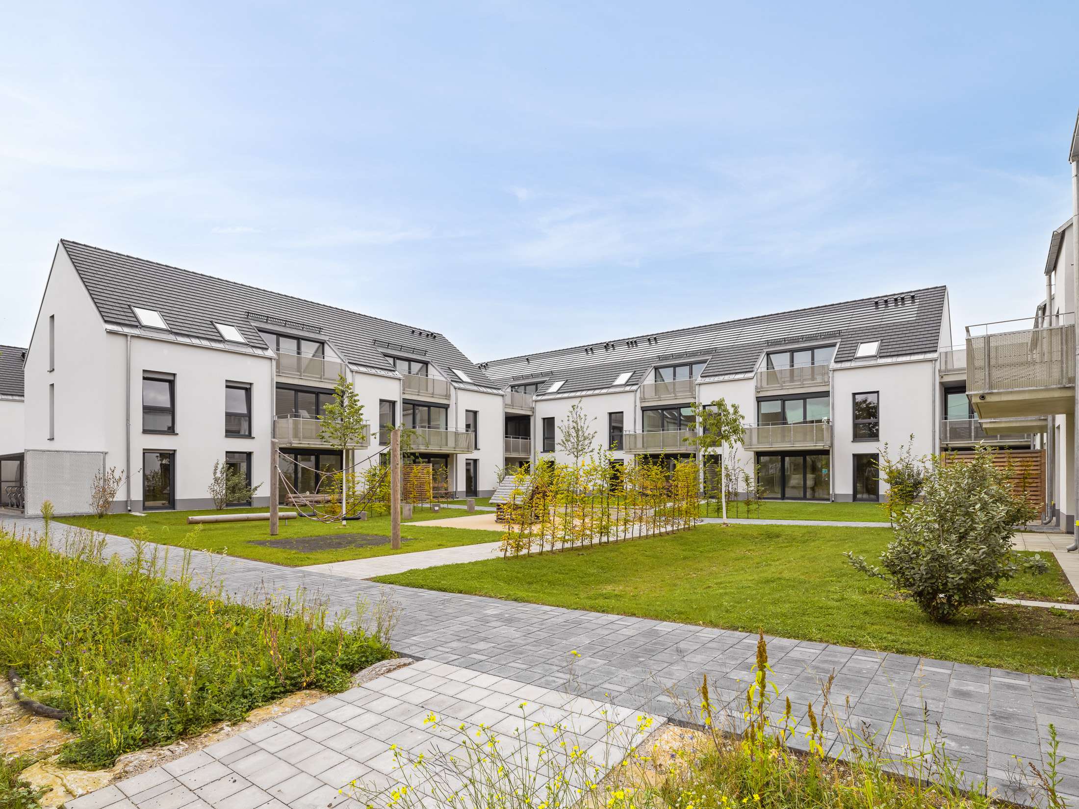 Mehrfamilienhausanlage für die VOLKSWOHNUNG GmbH in Karlsruhe-Durlach mit zweigeschossigen Hausgruppen und prägnanten Satteldächern, die sich um zwei begrünte, geschützte Innenhöfe organisieren; großzügige Verglasungen, Loggien und Dachgauben sowie eine flächenbündig integrierte Indach-Photovoltaikanlage prägen das architektonische Erscheinungsbild.