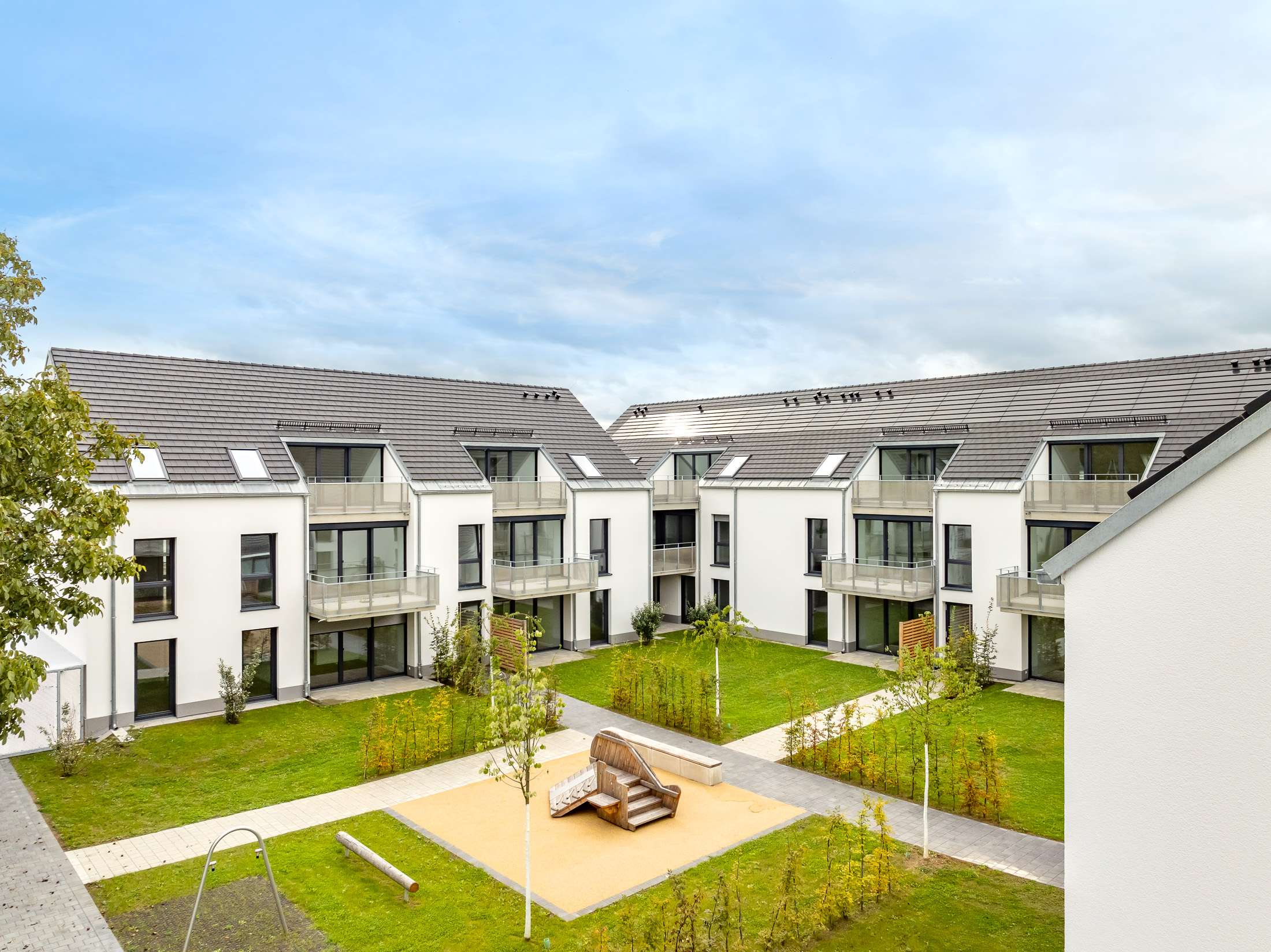 Mehrfamilienhausanlage für die VOLKSWOHNUNG GmbH in Karlsruhe-Durlach mit zweigeschossigen Hausgruppen und prägnanten Satteldächern, die sich um zwei begrünte, geschützte Innenhöfe organisieren; großzügige Verglasungen, Loggien und Dachgauben sowie eine flächenbündig integrierte Indach-Photovoltaikanlage prägen das architektonische Erscheinungsbild.