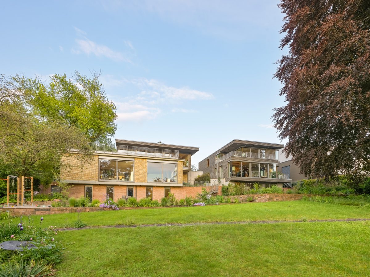 Architekturprojekt „Weiterbauen“ in Weinheim von Weber und Partner Architekten: Gesamtansicht der nachverdichteten Unternehmervilla aus den 1950er Jahren im parkartigen Hanggrundstück. Links der gekürzte Bestandsbau mit hellen Klinkern und Sandsteinsockel, rechts der moderne Erweiterungsbau aus Massivholz, verbunden durch eine gemeinsame Sandsteinstützmauer.