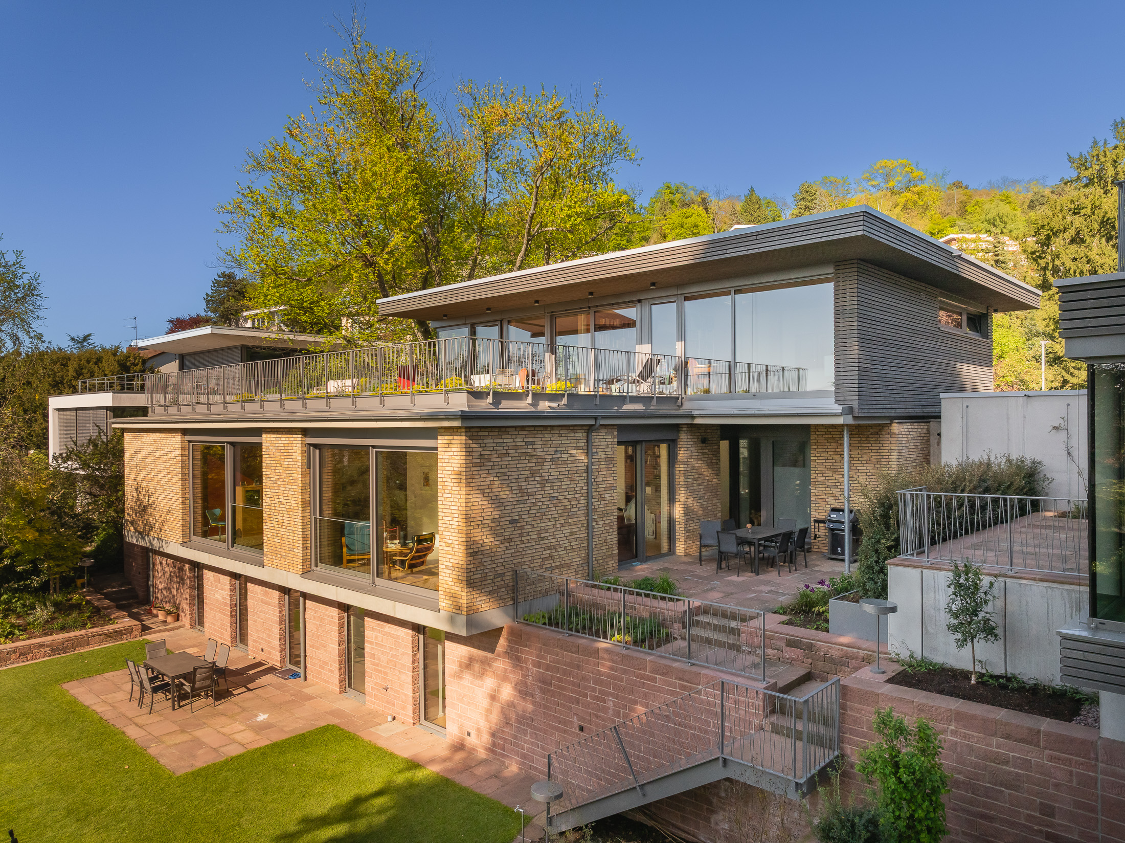 Architekturprojekt „Weiterbauen“ in Weinheim von Weber und Partner Architekten: Gesamtansicht der nachverdichteten Unternehmervilla aus den 1950er Jahren im parkartigen Hanggrundstück. Der gekürzte Bestandsbau mit hellen Klinkern und Holzaufstockung verbindet sich durch den Sandsteinsockel mit dem modernen Erweiterungsbau aus Massivholz.