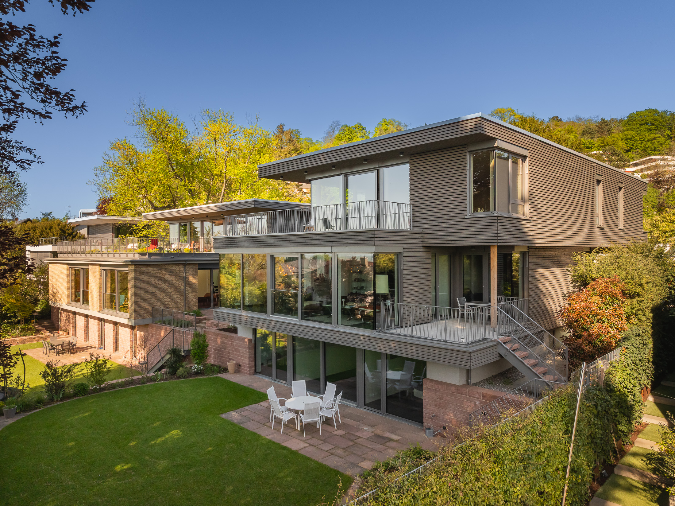 Architekturprojekt „Weiterbauen“ in Weinheim von Weber und Partner Architekten: Gesamtansicht der nachverdichteten Unternehmervilla aus den 1950er Jahren im parkartigen Hanggrundstück. Der gekürzte Bestandsbau mit hellen Klinkern und Holzaufstockung verbindet sich durch den Sandsteinsockel mit dem modernen Erweiterungsbau aus Massivholz.
