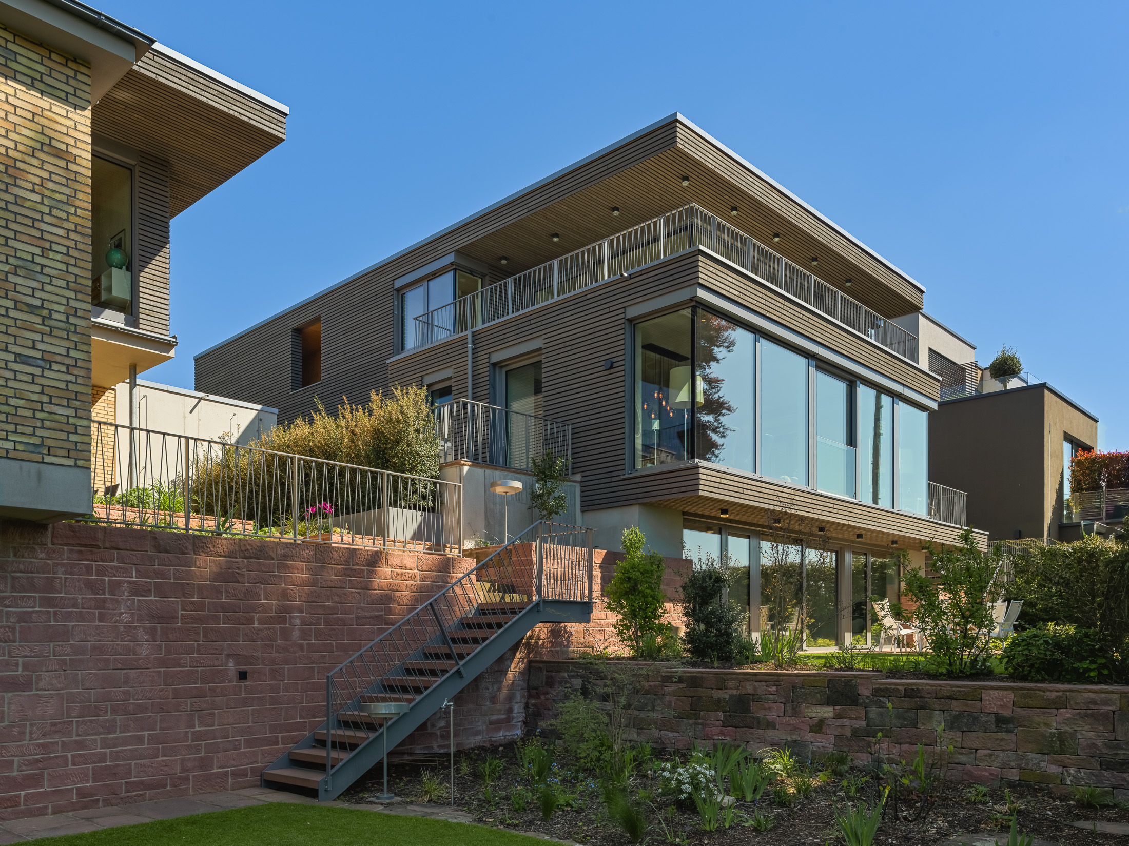 Architekturprojekt „Weiterbauen“ in Weinheim von Weber und Partner Architekten: Gesamtansicht der nachverdichteten Unternehmervilla aus den 1950er Jahren im parkartigen Hanggrundstück. Der gekürzte Bestandsbau mit hellen Klinkern und Holzaufstockung verbindet sich durch den Sandsteinsockel mit dem modernen Erweiterungsbau aus Massivholz.