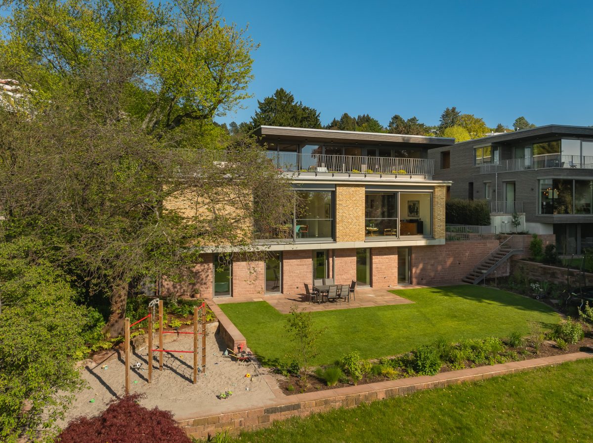 Architekturprojekt „Weiterbauen“ in Weinheim von Weber und Partner Architekten: Gesamtansicht der nachverdichteten Unternehmervilla aus den 1950er Jahren im parkartigen Hanggrundstück. Links der gekürzte Bestandsbau mit hellen Klinkern und Sandsteinsockel, rechts der moderne Erweiterungsbau aus Massivholz, verbunden durch eine gemeinsame Sandsteinstützmauer.