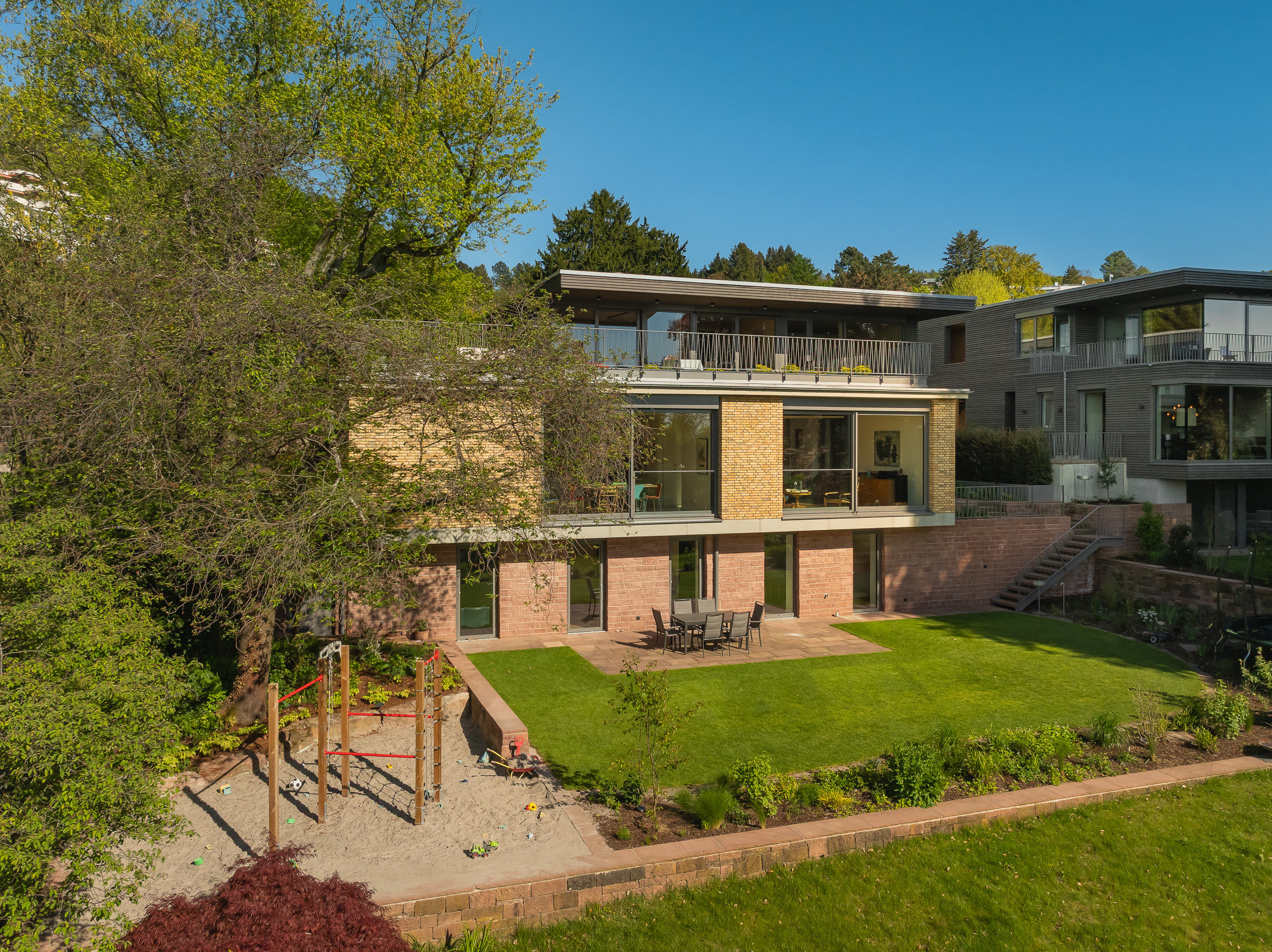 Architekturprojekt „Weiterbauen“ in Weinheim von Weber und Partner Architekten: Gesamtansicht der nachverdichteten Unternehmervilla aus den 1950er Jahren im parkartigen Hanggrundstück. Links der gekürzte Bestandsbau mit hellen Klinkern und Sandsteinsockel, rechts der moderne Erweiterungsbau aus Massivholz, verbunden durch eine gemeinsame Sandsteinstützmauer.